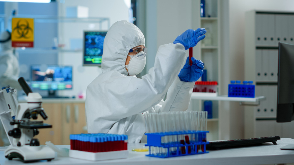 A laboratory technician in full protective gear, including a hazmat suit, face mask, goggles, and gloves, examines a blood sample tube in a modern microbiology laboratory. Test tubes, laboratory equipment, and monitoring screens are visible, representing microbiological testing and quality analysis services in Qatar.