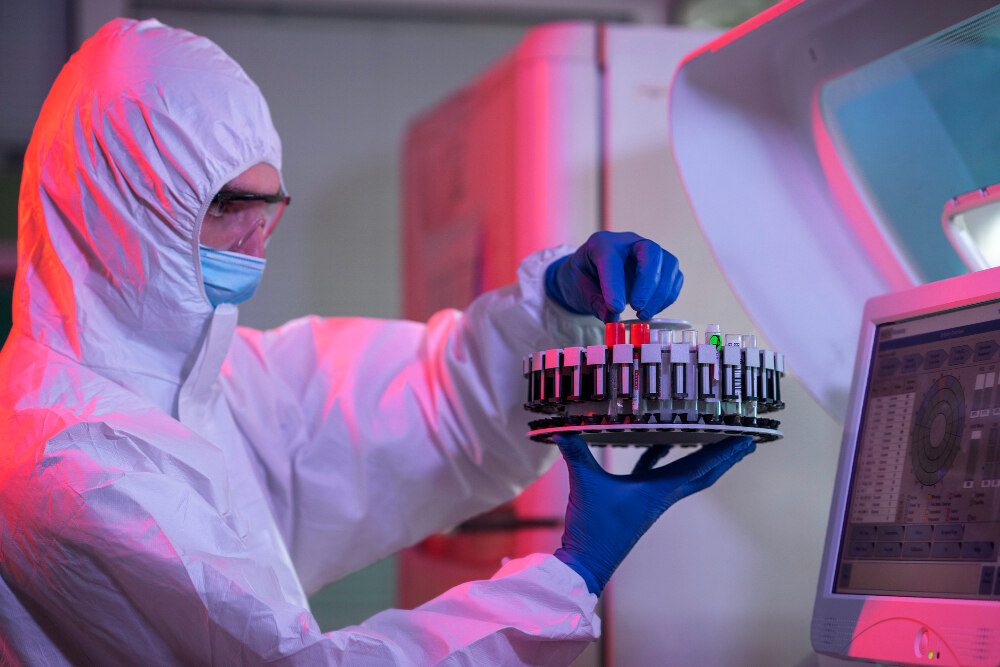 Laboratory scientist wearing protective clothing, face mask, and gloves handling sample tubes in an automated microbiological testing system, conducting laboratory analysis in a controlled environment.